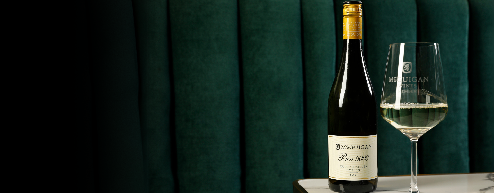 McGuigan Bin 900 Semillon on a marble tale with a velvet green sofa in the background. an etched McGuigan Wines wine glass stands beside full of white wine.