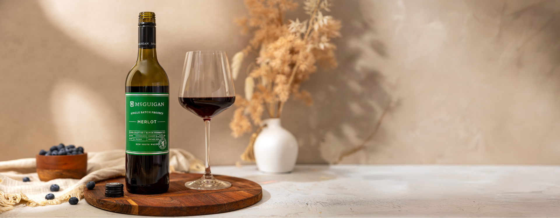 McGuigan Single Batch Merlot wine against a beige textured backdrop and a bowl of blueberries