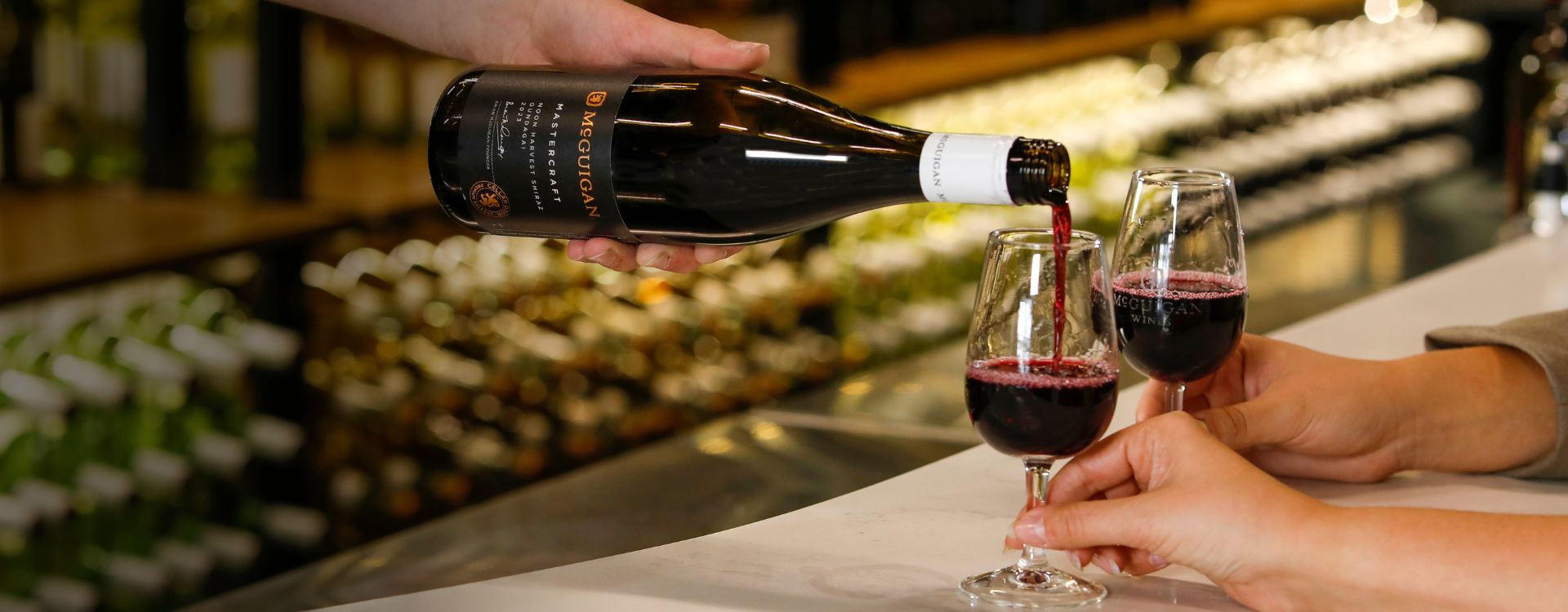 McGuigan red wine bottle being poured into two glasses held by hands across the bar.